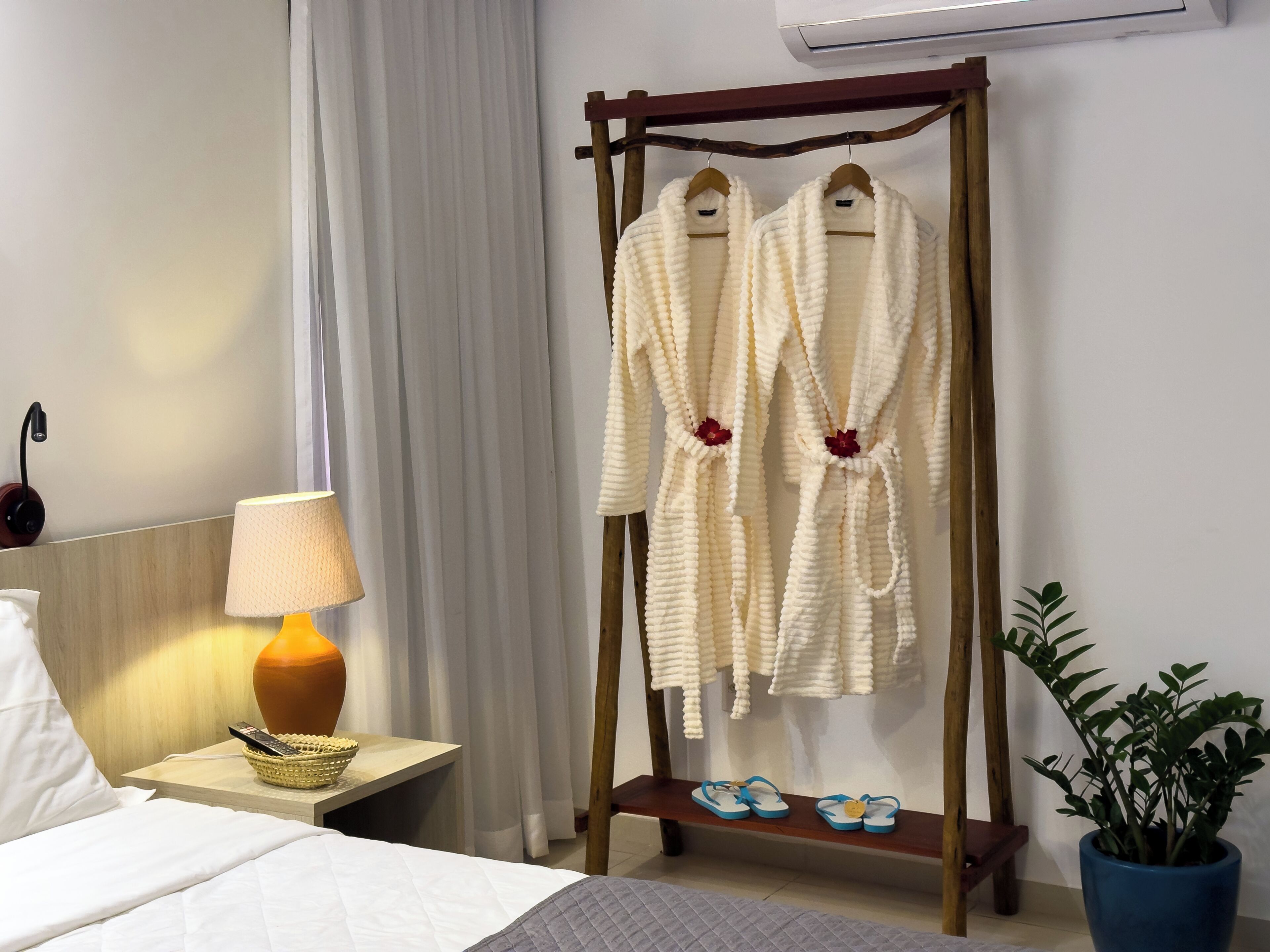 A hotel room with a bed, a bedside lamp, a wooden rack with robes, a pair of slippers, a potted plant, and a wall-mounted air conditioner.