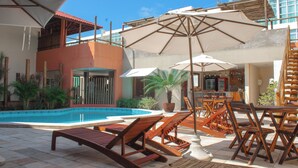 A poolside area with lounge chairs and umbrellas, a pool, and a building with a red roof and multiple levels. - Vivá Barra Hotel Pousada (Barra de São Miguel)