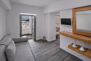 A modern living room with a grey sofa, a wooden TV stand, and a balcony with a white railing. - Thireas Hotel (Santorini)