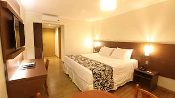 Quarto de hotel com cama, mesas de cabeceira, escrivaninha, cadeira e televisão.