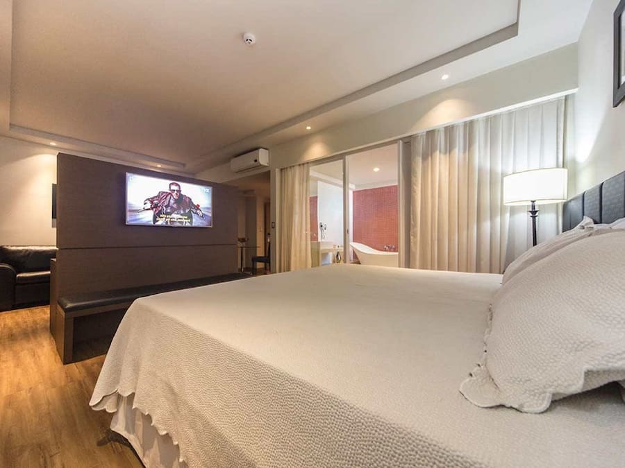 Una habitación de hotel moderna con una cama grande, un televisor de pantalla plana montado en la pared, una zona de estar con sofá y un ventanal con cortinas.