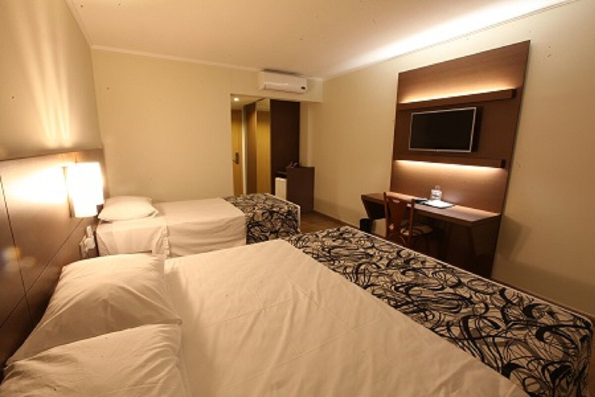 A hotel room with two beds, a desk, a chair, a television, and an air conditioning unit.
