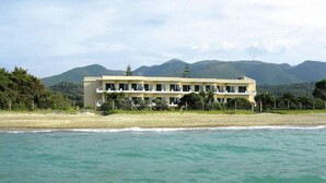 A two-story hotel with a flat roof, located on a beach with a view of mountains in the background. - Akti Anastasia (Corfu)