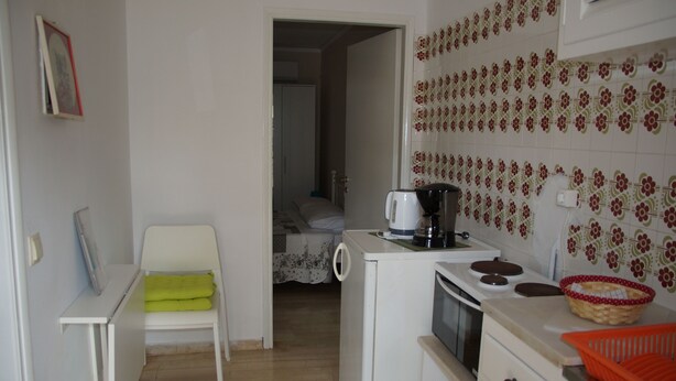 A compact kitchen with a small stove, a coffee maker, and a sink. A chair with a green towel is placed near the entrance.