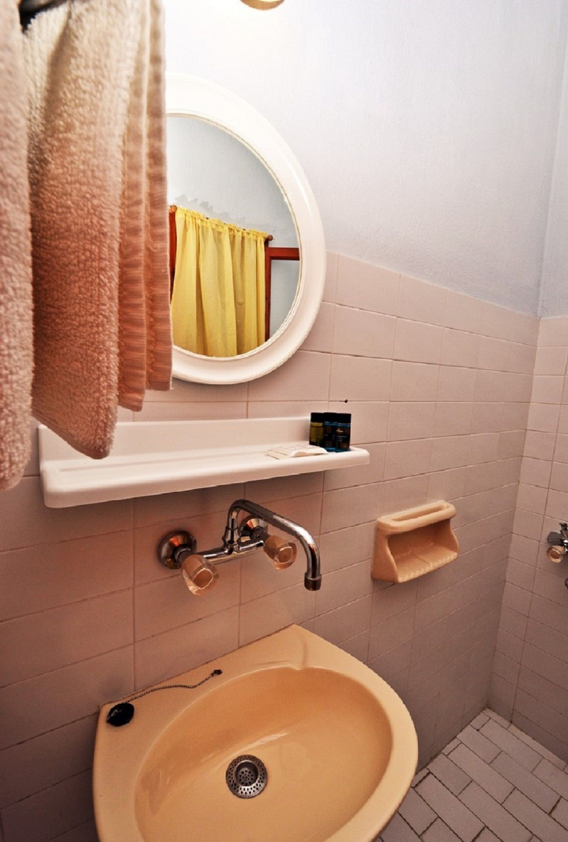 A bathroom with a beige sink, a mirror, a towel rack, and a shelf with toiletries.