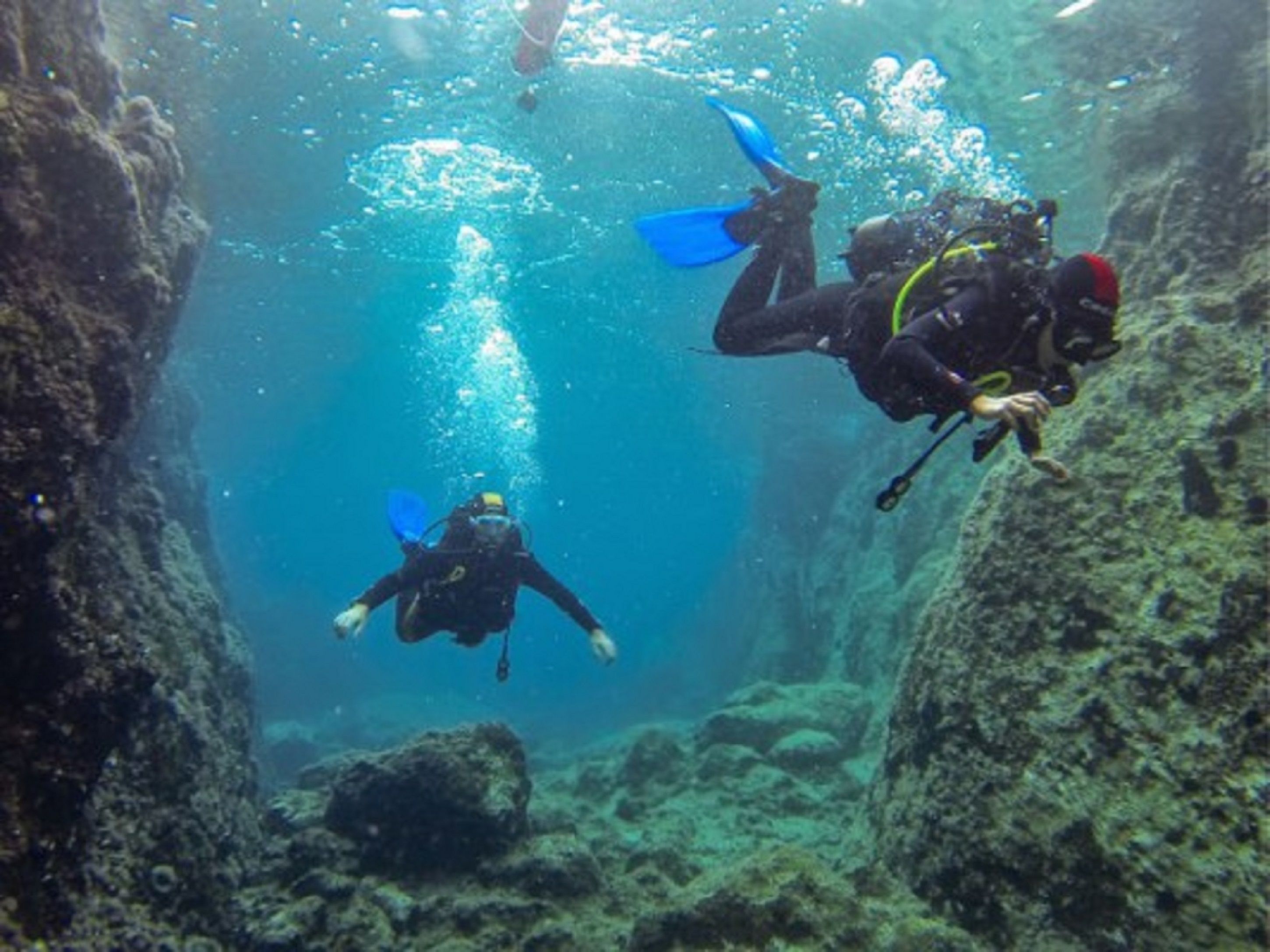 Three divers in a narrow underwater passage with rocky walls and marine life.