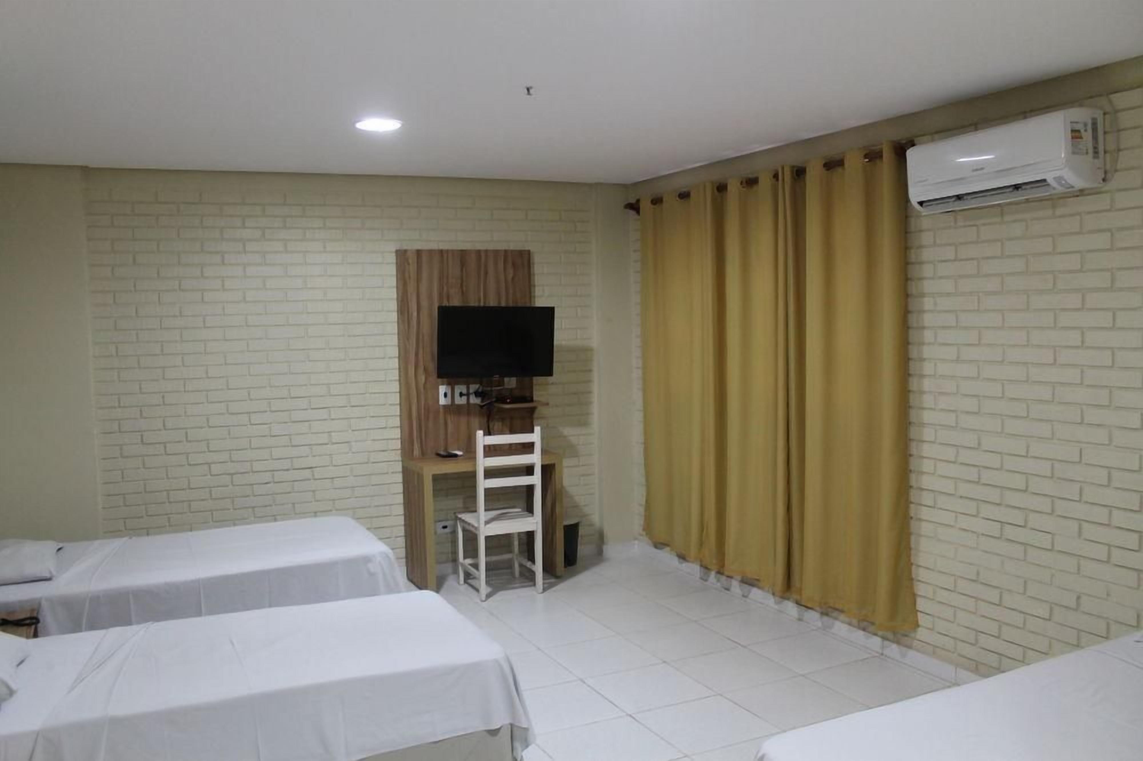 Quarto de hotel com duas camas, televisão, escrivaninha e cadeira, e aparelho de ar condicionado.