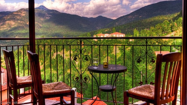 A balcony with two wooden chairs and a small table, overlooking a mountainous landscape with a building in the distance. - Ta Petrina (Nafpaktos)