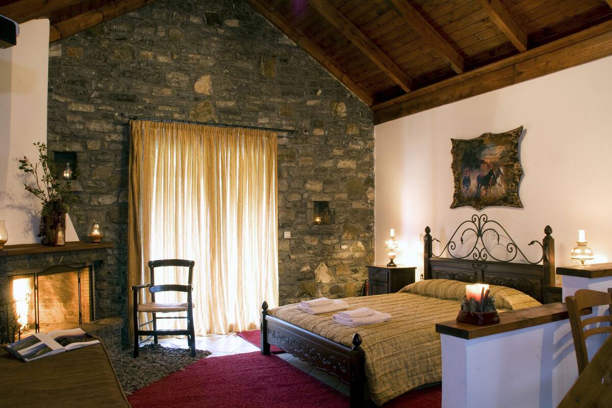 A bedroom with a stone wall, a bed with a wooden headboard, a chair, a bedside table with a lamp, and a fireplace.