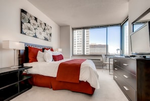A modern hotel room with a large bed, a desk, and a view of the city through large windows.