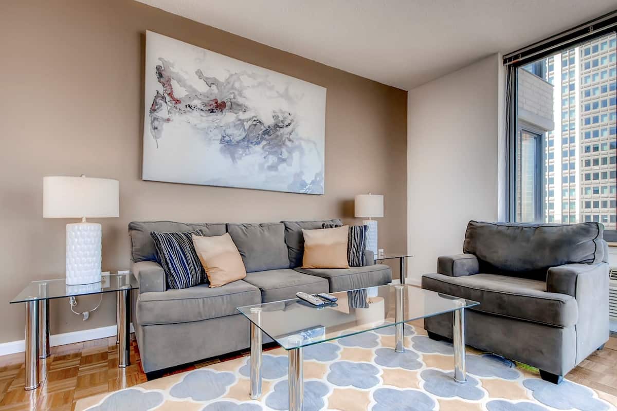 Un salon moderne avec un canapé gris, une table basse en verre et un fauteuil gris. Deux lampes sont posées sur des tables d’appoint, et un grand tableau abstrait orne le mur.