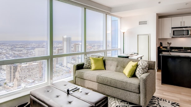 A modern living room with a large window, a grey sofa, a small table, and a kitchen area. - Global Luxury Suites at Columbus (Jersey City)