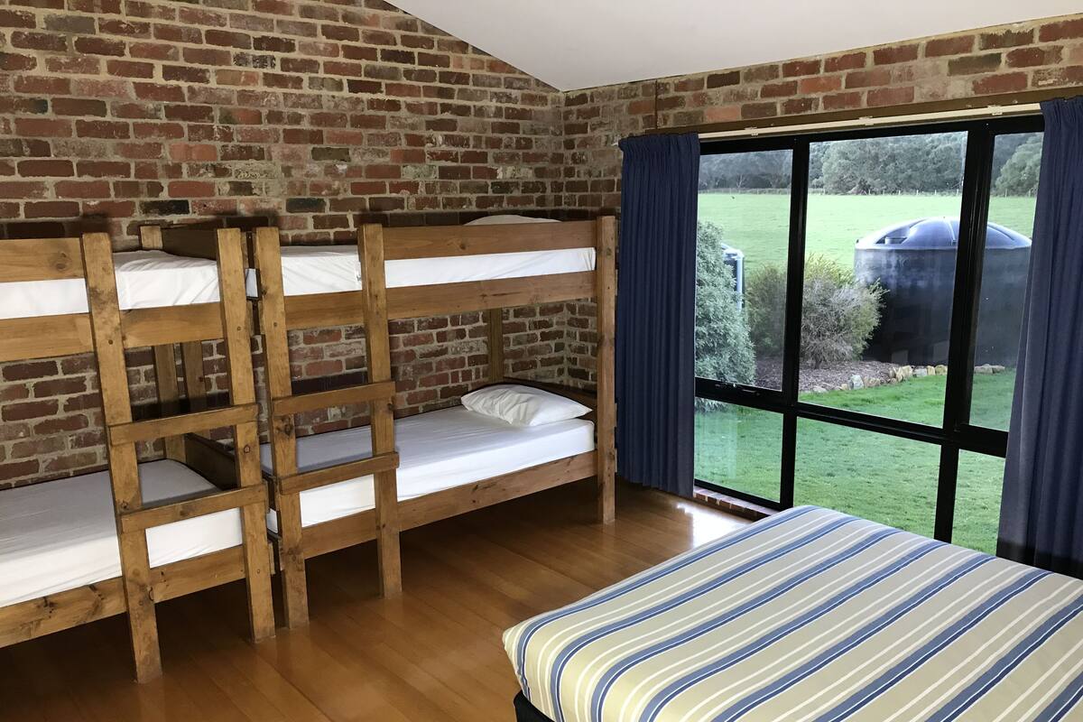 A room with three bunk beds and a single bed, brick walls, and a large window with a view of a green landscape.