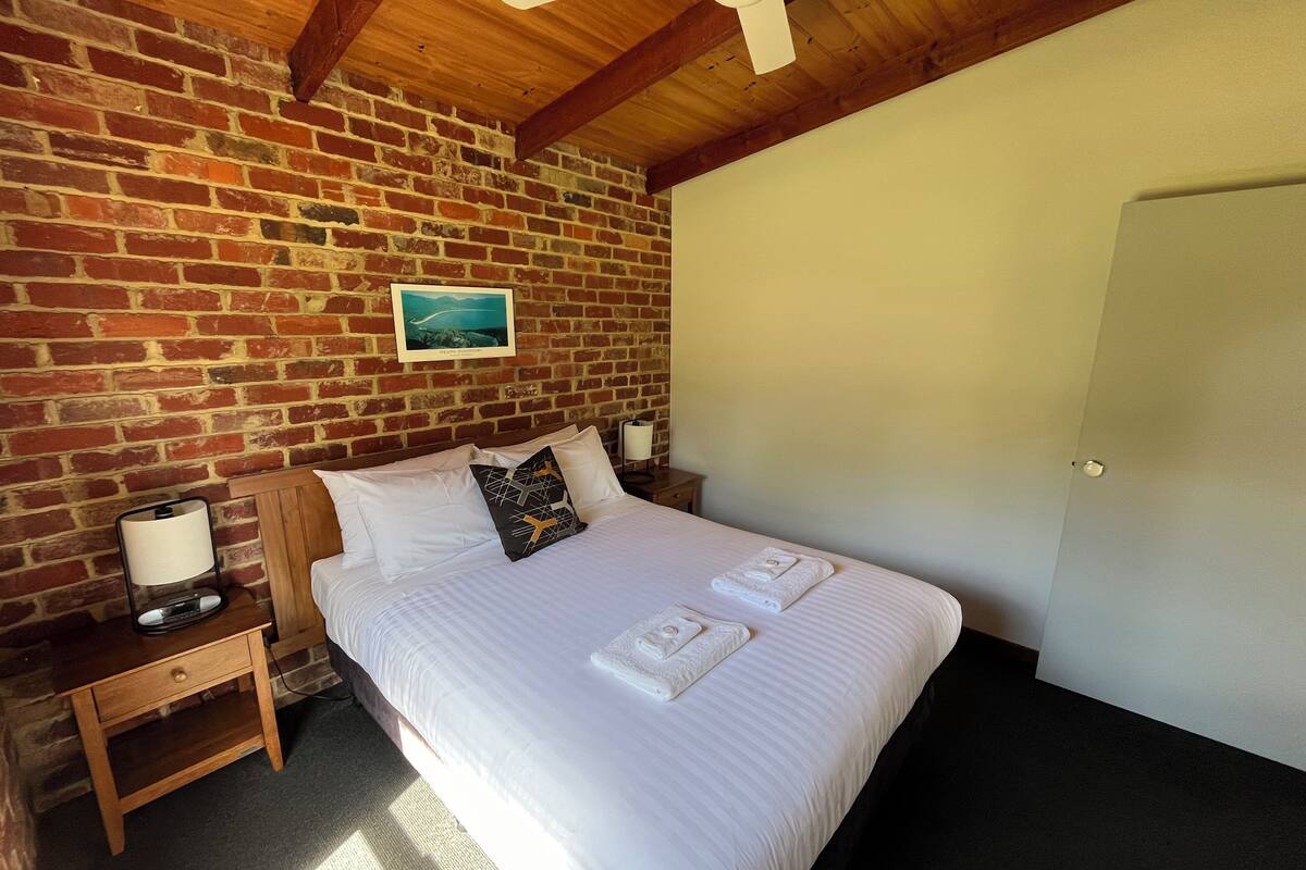 A room with a brick wall, a bed with white linens, a nightstand with a lamp, and a ceiling fan.