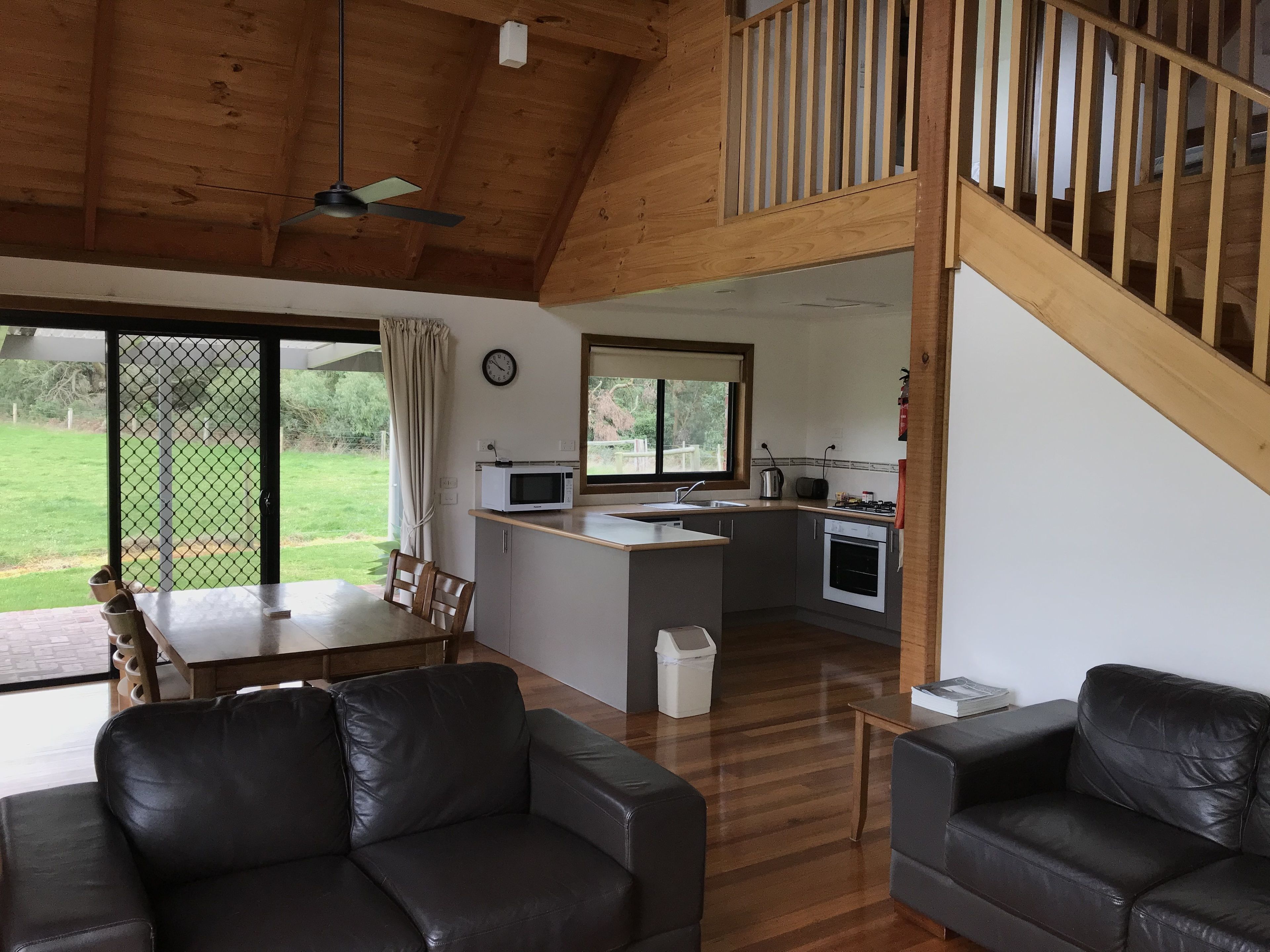 Ein geräumiger Wohnbereich mit Holzboden, einem Esstisch und zwei Ledersofas. Im Hintergrund ist eine Küche mit Mikrowelle und Herd erkennbar.
