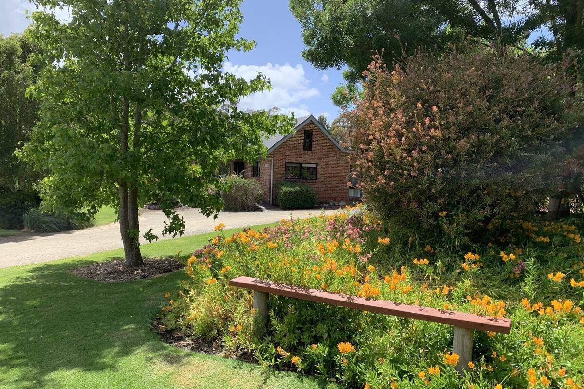 A brick building with a sloped roof, surrounded by a well-maintained garden with various flowering plants and a wooden bench.