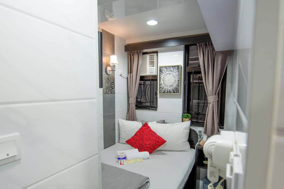 Une chambre d’hôtel de taille réduite, comprenant un lit simple, une table de chevet, un petit bureau et une fenêtre avec des rideaux.