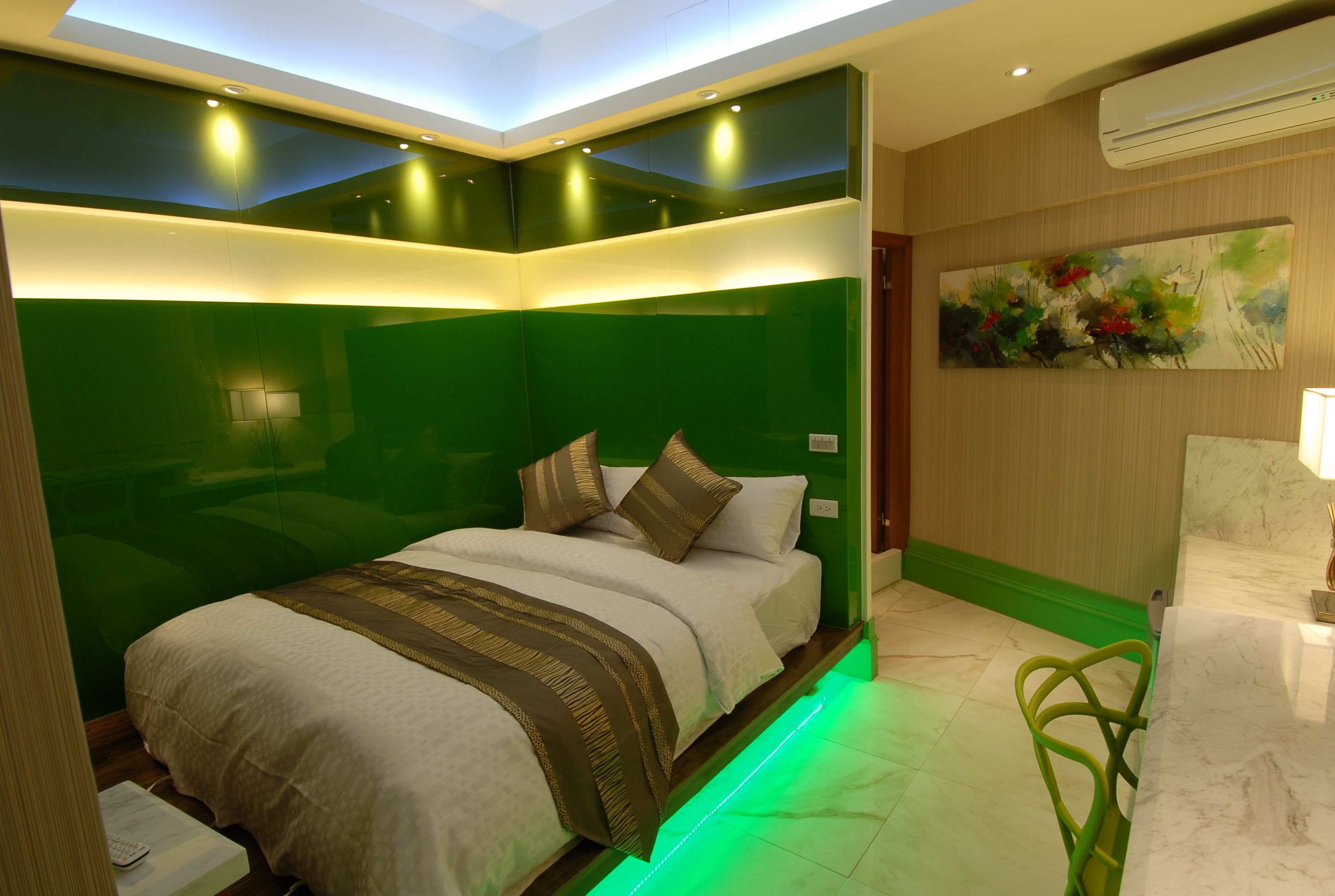 Quarto de hotel moderno com uma parede verde de destaque, uma cama com colcha estampada, uma cadeira verde e um quadro na parede.