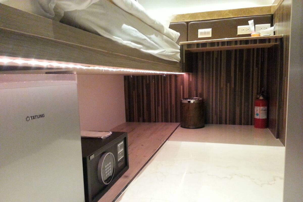A compact room with a bed, a minibar, a trash bin, and a fire extinguisher.