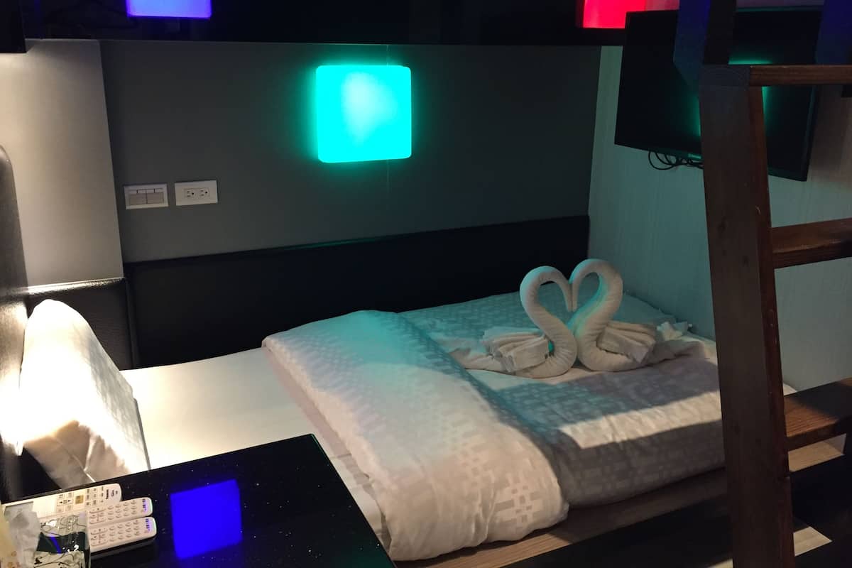 A hotel room with a bed, a bedside table with a lamp, a wall-mounted TV, and a neon sign.