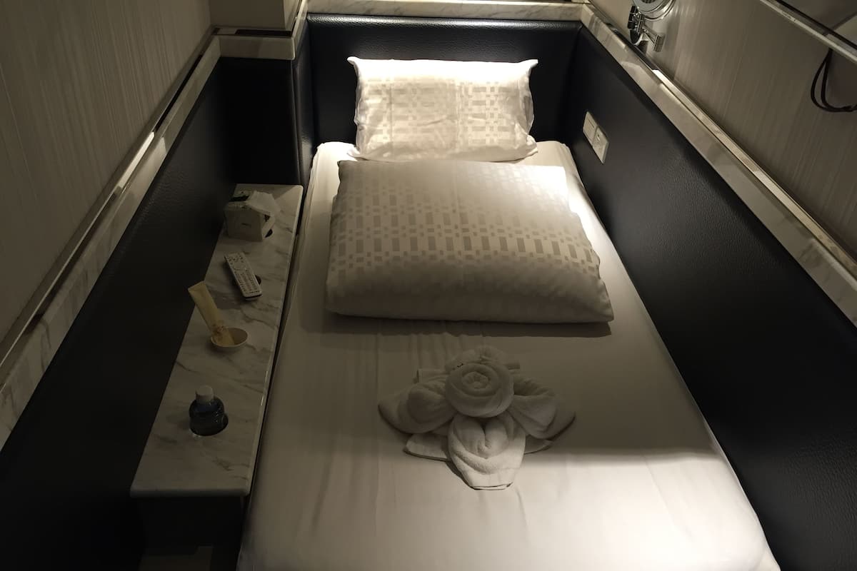 A single bed with a white pillow and a towel with a floral design. There is a remote control and a small bottle on the nightstand.
