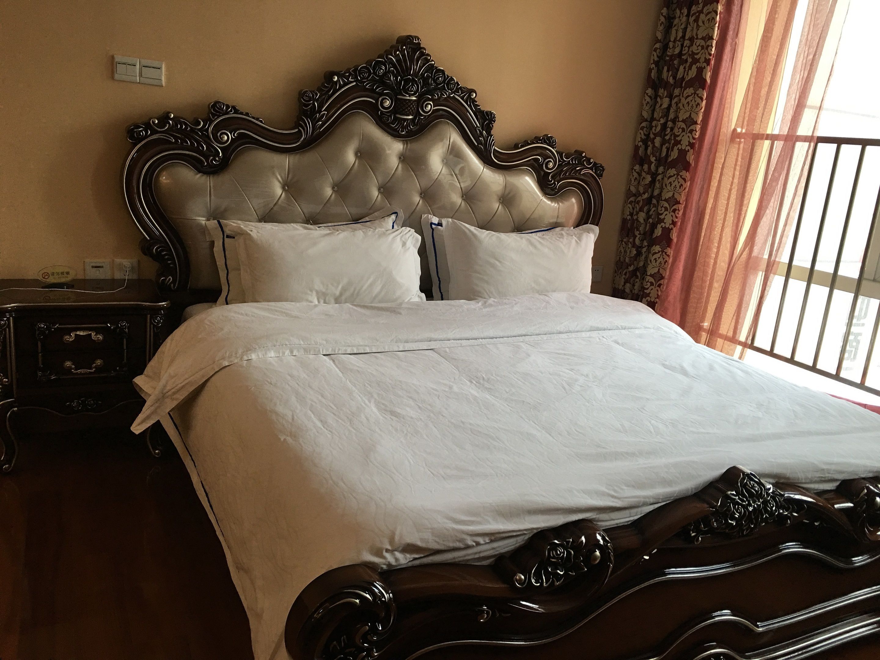 Una cama bien hecha con cabecera capitonada, una mesita de noche de madera con cajones y una ventana con cortinas traslúcidas.
