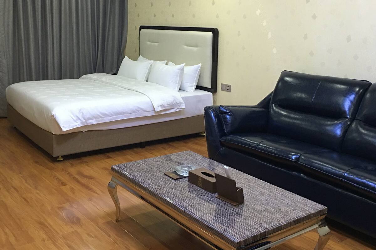 Habitación de hotel con una cama, un sofá de cuero negro y una mesita con una caja de pañuelos y un control remoto.