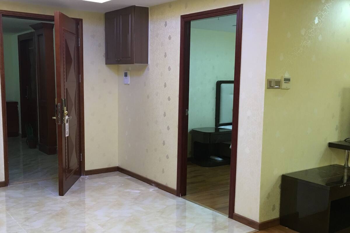 Una habitación con piso de madera, una puerta que da a otra habitación, un mueble pequeño y un termostato en la pared.