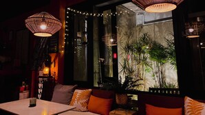 Interior de un restaurante tenuemente iluminado con una zona de estar acogedora, luces colgantes y un amplio ventanal con vistas a la vegetación.