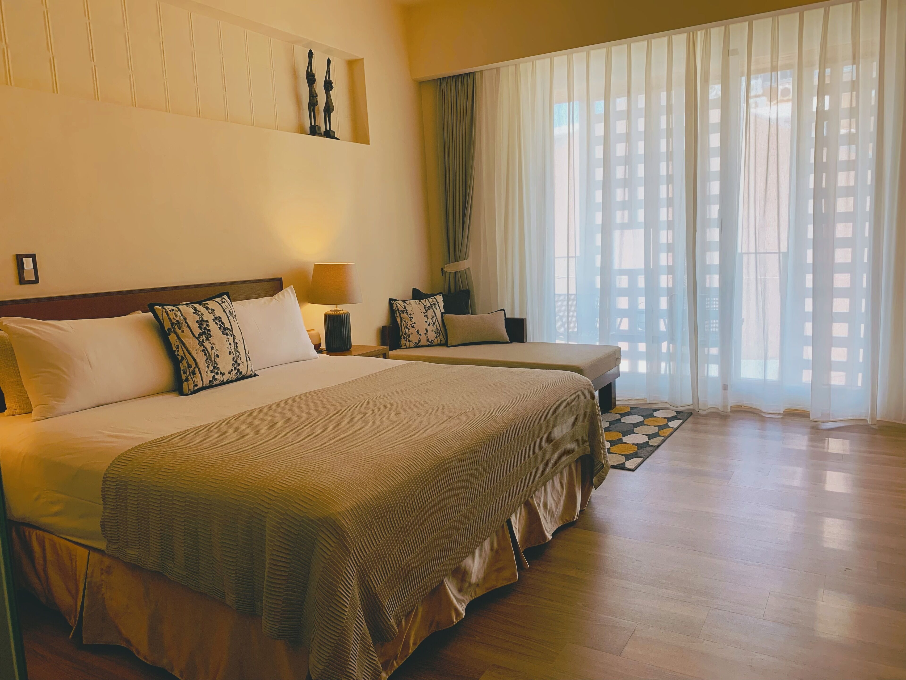 A hotel room with two beds, a large window with sheer curtains, a wooden floor, and a wall-mounted artwork.