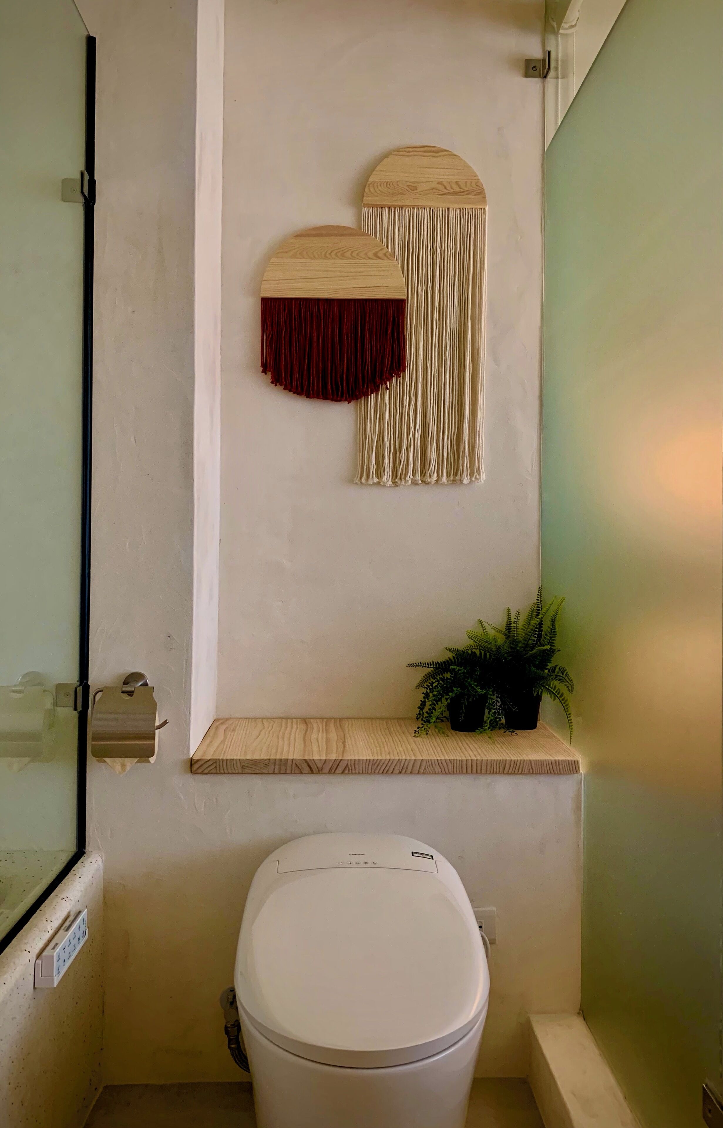 Un baño pequeño con inodoro, un estante de madera con una planta y un adorno de macramé en la pared.