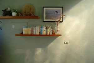 Una estantería con libros y un jarrón con flores secas, un cuadro enmarcado en la pared y un enchufe.