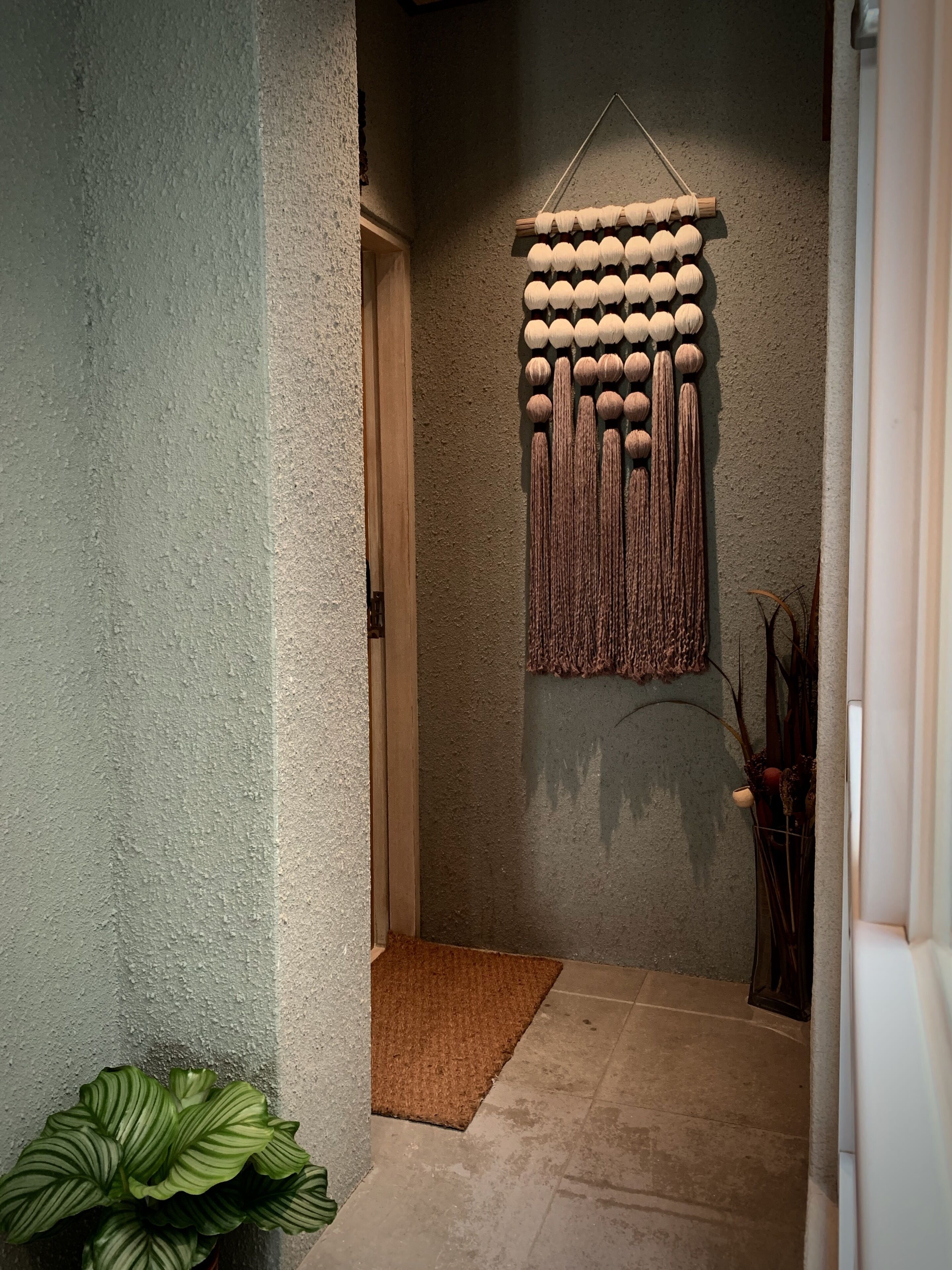 Un couloir avec un mur à texture, une décoration murale en macramé, une plante en pot et un paillasson.