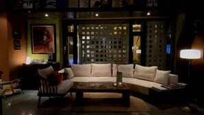 Una sala moderna con un sofá seccional, una mesa de centro y una lámpara. Se observa un separador decorativo detrás del sofá y un cuadro en la pared.