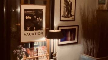 Una habitación con un escritorio, una lámpara, un cuadro enmarcado que dice 'VACATION' y un jarrón grande con flores.