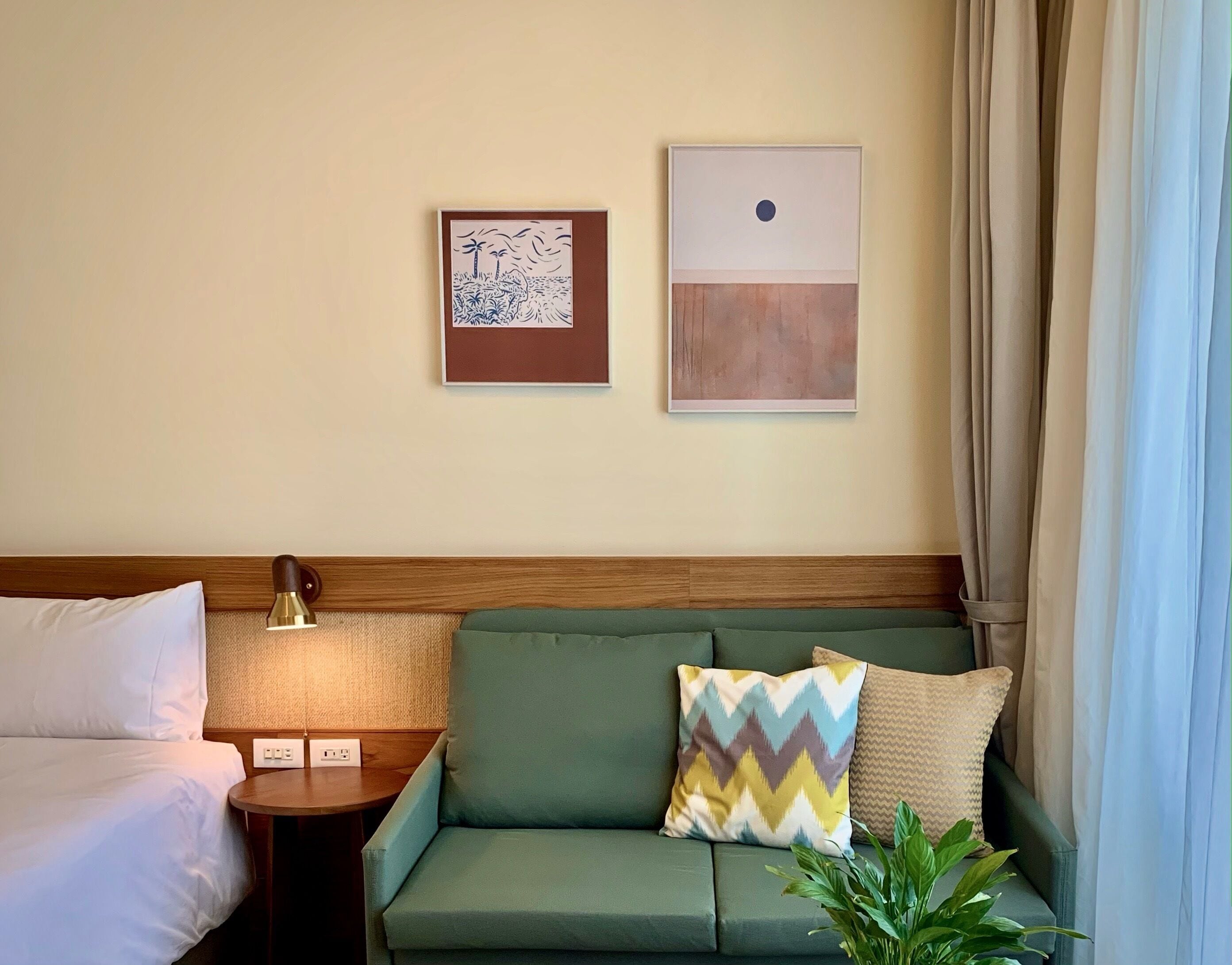 A hotel room with a bed, a green sofa with patterned cushions, a small table with a lamp, and two wall art pieces.