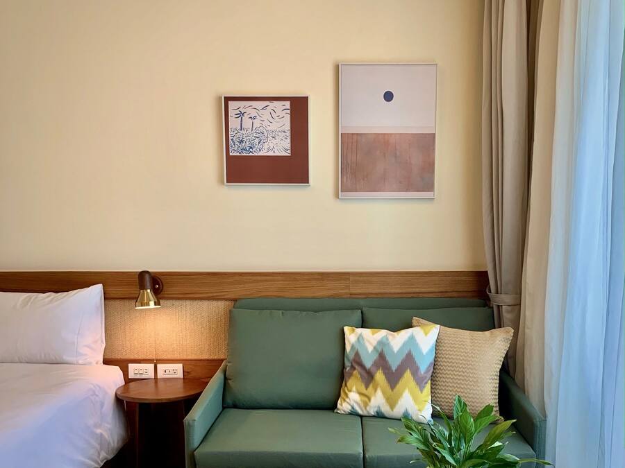 Ein Hotelzimmer mit einem Bett, einem grünen Sofa mit gemusterten Kissen, einem kleinen Tisch mit einer Lampe und zwei Wandbildern.