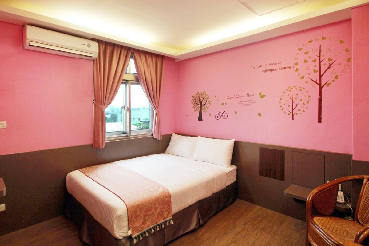 Una habitación de hotel con cama, ventana con cortinas, pared con un vinilo de árbol y una silla.