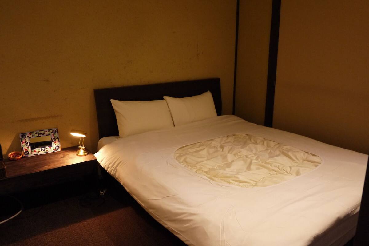 Un lit bien fait dans une chambre dont la tête de lit est de couleur sombre, avec une table de chevet sur laquelle se trouve une lampe, et un tableau encadré accroché au mur.