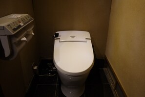 Un inodoro blanco estándar en un baño pequeño y poco iluminado, con un panel de control a la izquierda.