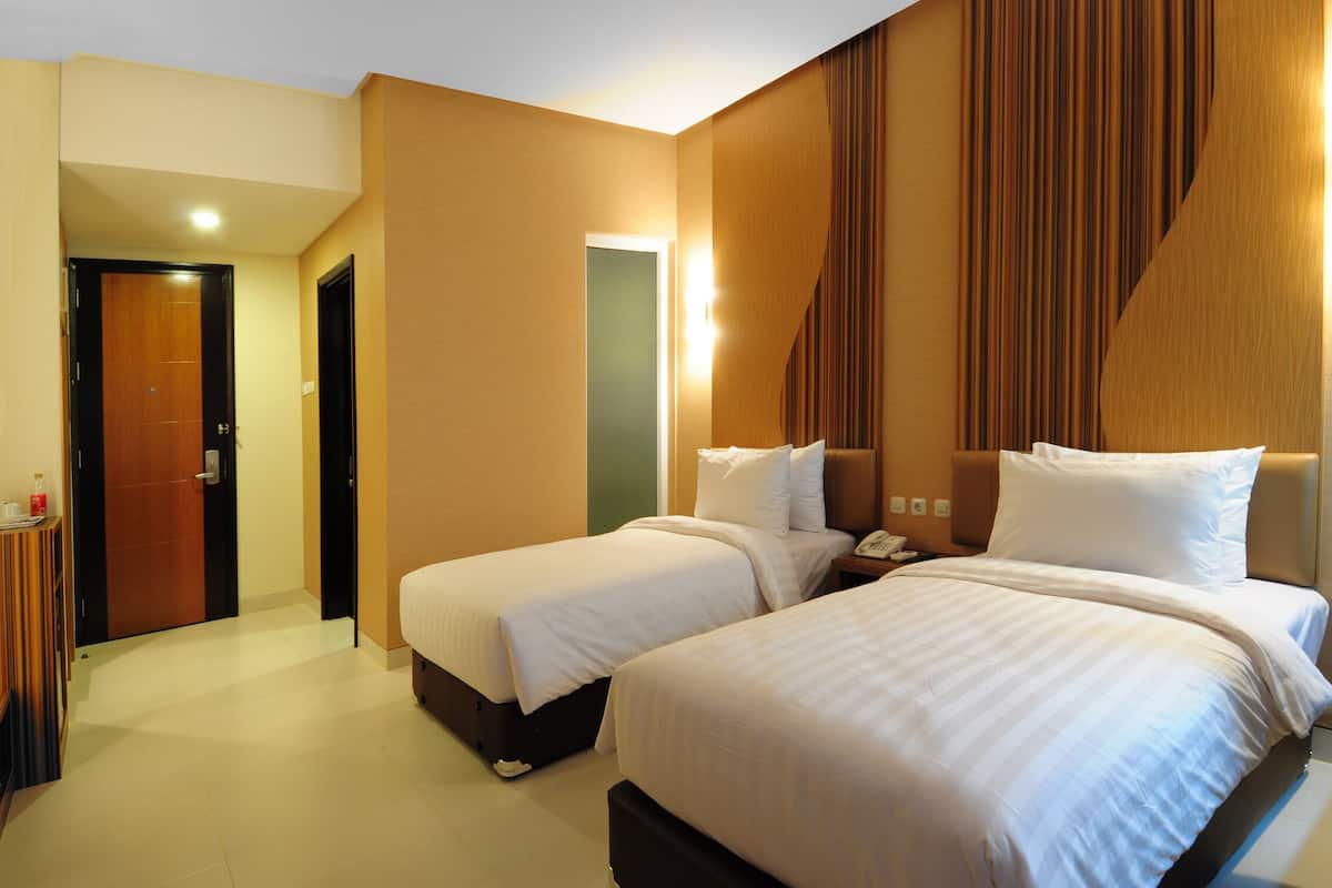 A hotel room with two beds, a bedside table, a nightstand, and a wall-mounted lamp.