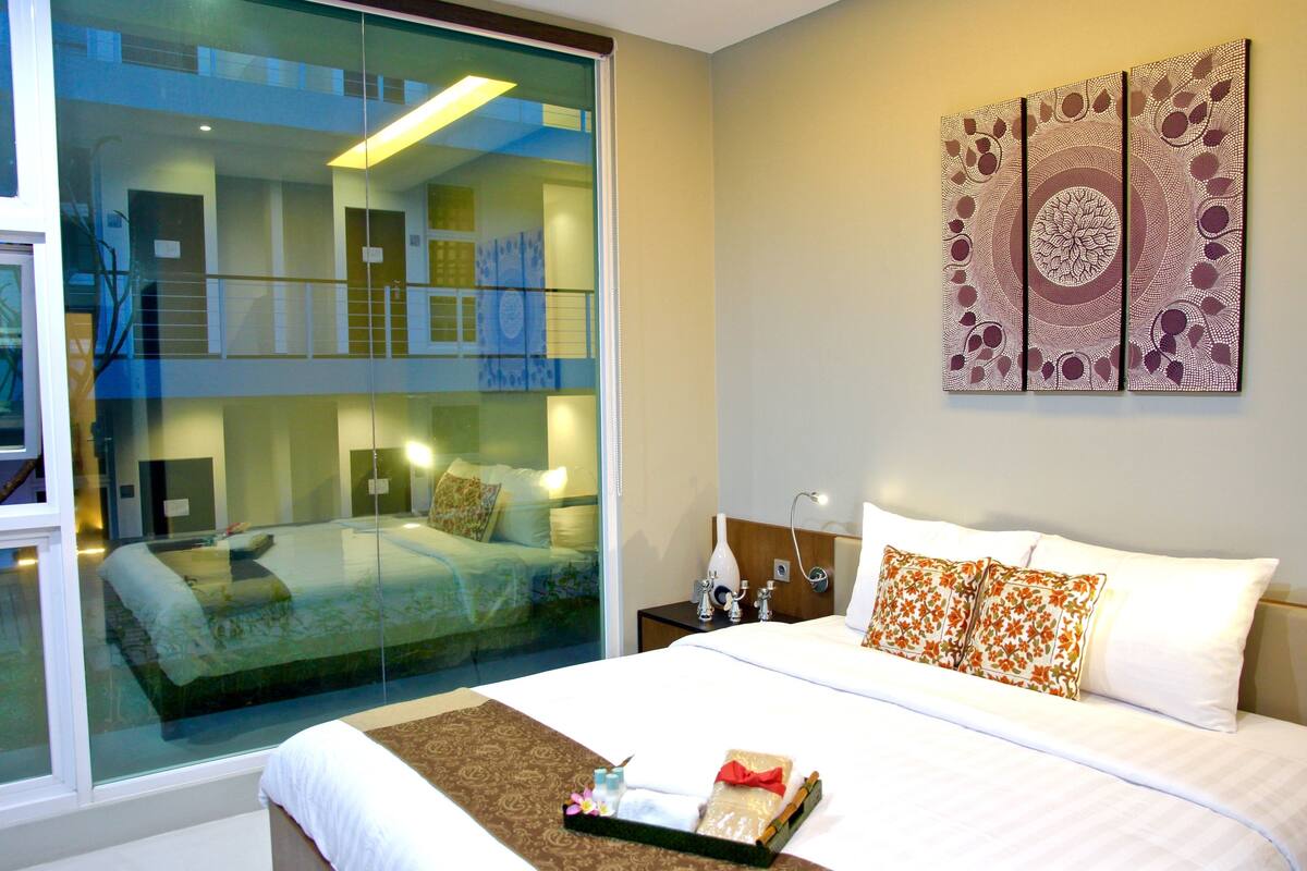 A hotel room with a large bed, a glass wall, and a decorative wall piece.