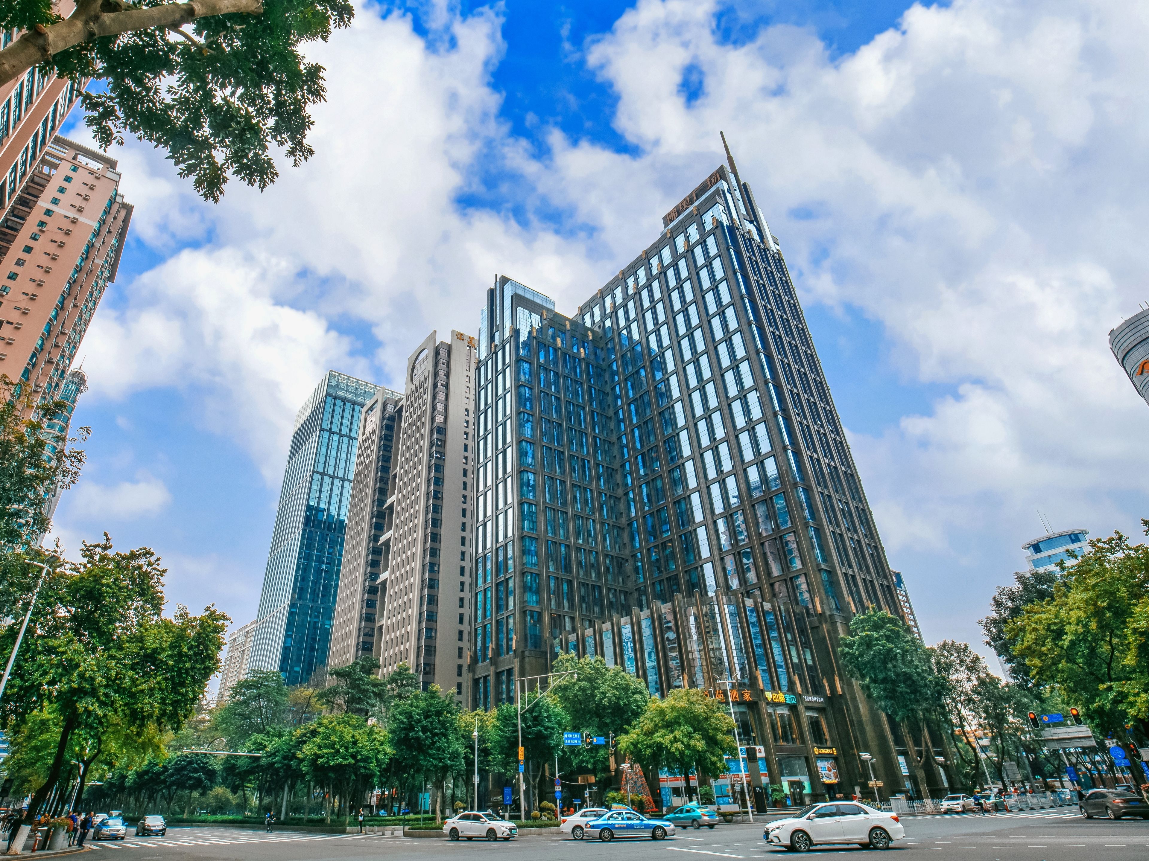 A modern high-rise building with a glass facade, surrounded by trees and other buildings.