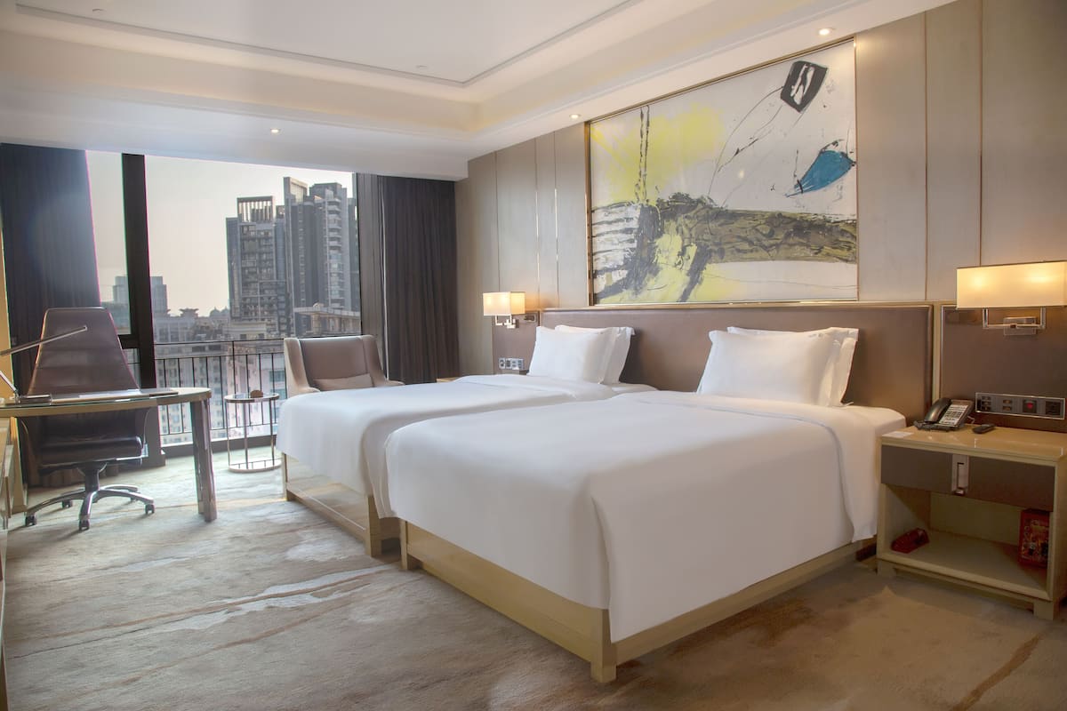 Une chambre d’hôtel avec deux lits, un bureau, une chaise, une table de chevet et une grande fenêtre donnant sur la ville.