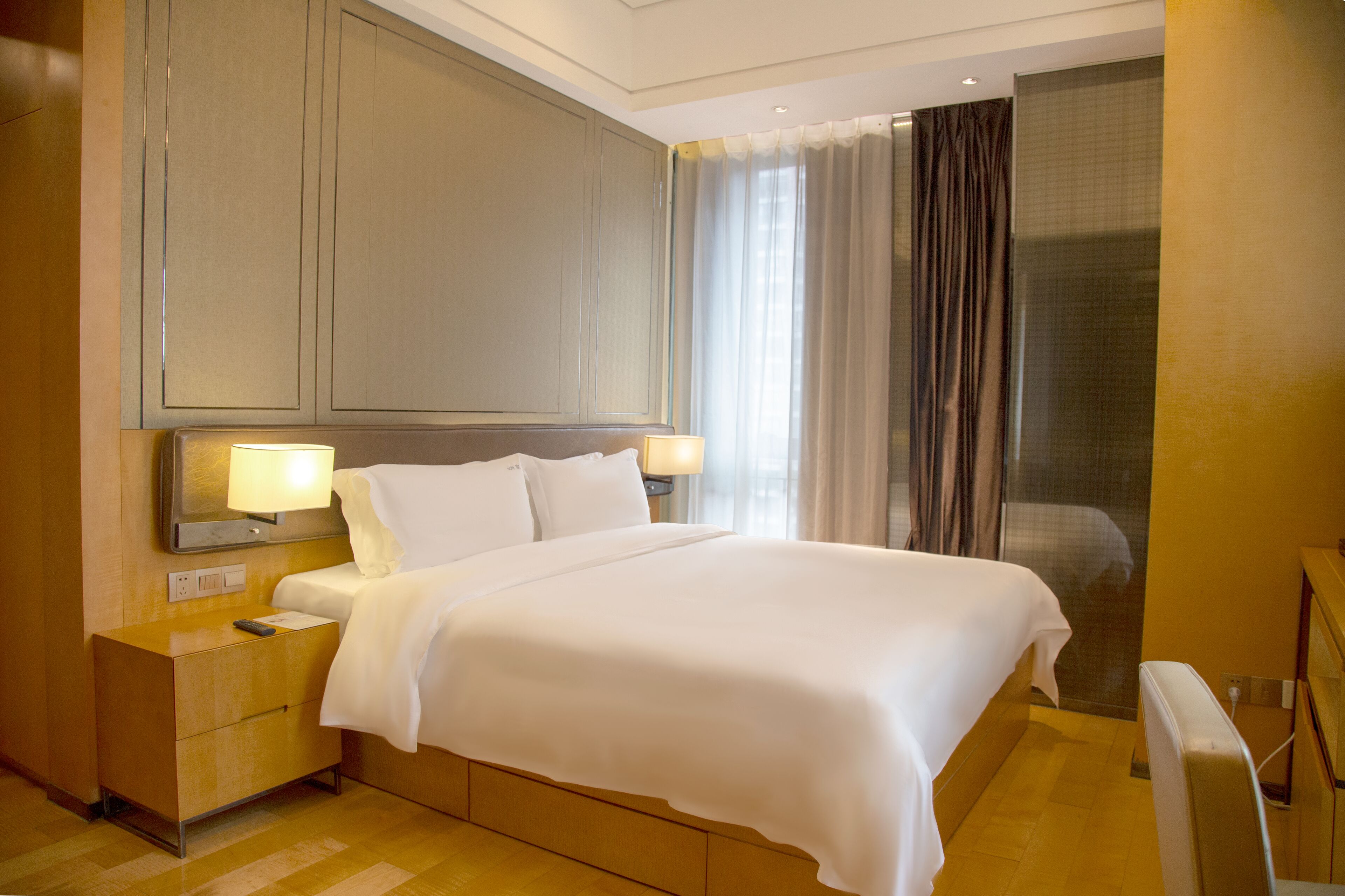 A hotel room with a large bed, bedside lamps, a nightstand, and a view of the outside through a window with curtains.