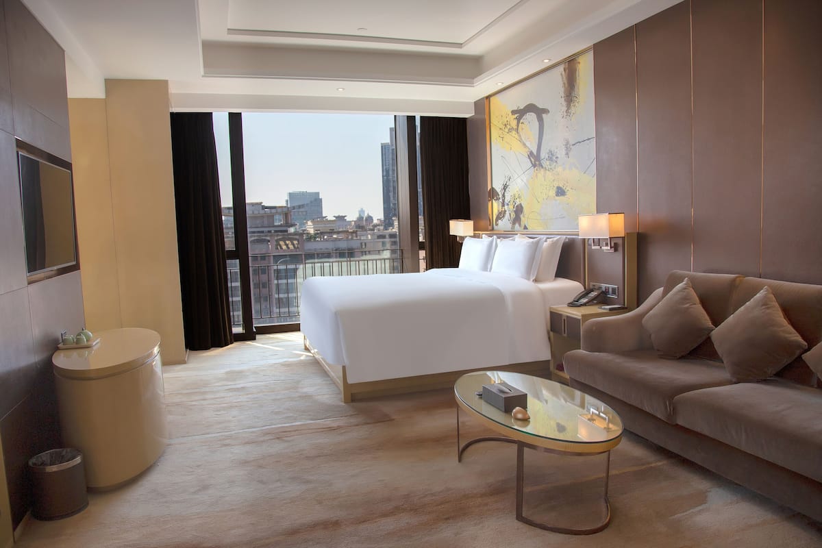 Une chambre d’hôtel moderne dotée d’un grand lit, d’un canapé, d’une table basse en verre, d’une table de chevet avec des lampes et offrant une vue sur la ville.