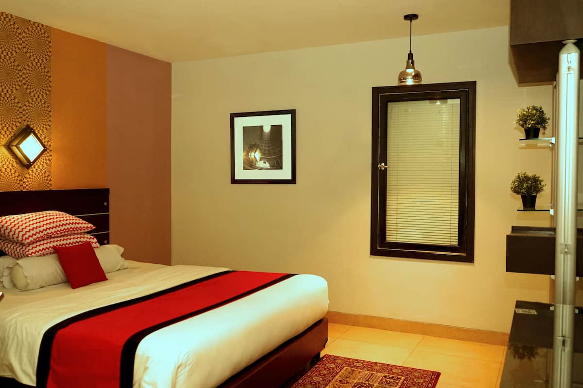 A bedroom with a bed, a window with blinds, a framed picture on the wall, and a small potted plant on a shelf.
