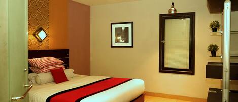 A bedroom with a bed, a window with blinds, a framed picture on the wall, and a small potted plant on a shelf.