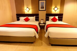 A hotel room with two beds, each with white bedding and red accents, a nightstand with a phone and a framed picture above it, and a wall-mounted light fixture. - Citi M Hotel (Jakarta)