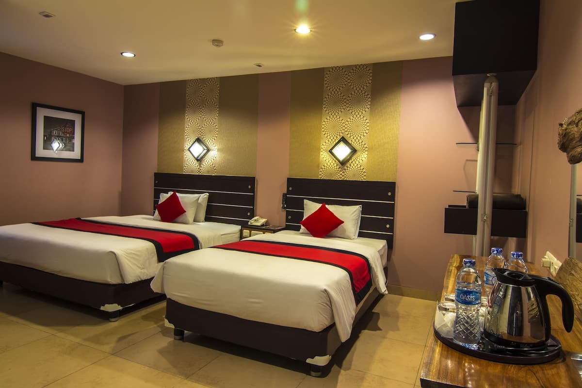 A hotel room with two beds, a wooden table with a kettle and water bottles, and a wall-mounted TV.